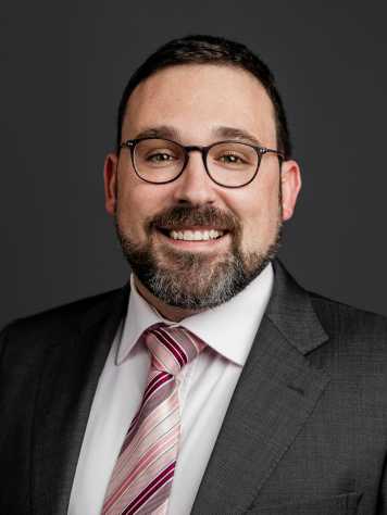 A formal portrait photo of a man with short brown hair and a beard wearing glasses.