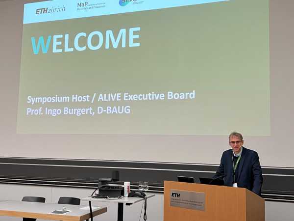 a man at the speaker's desk in front of a big screen saying welcome